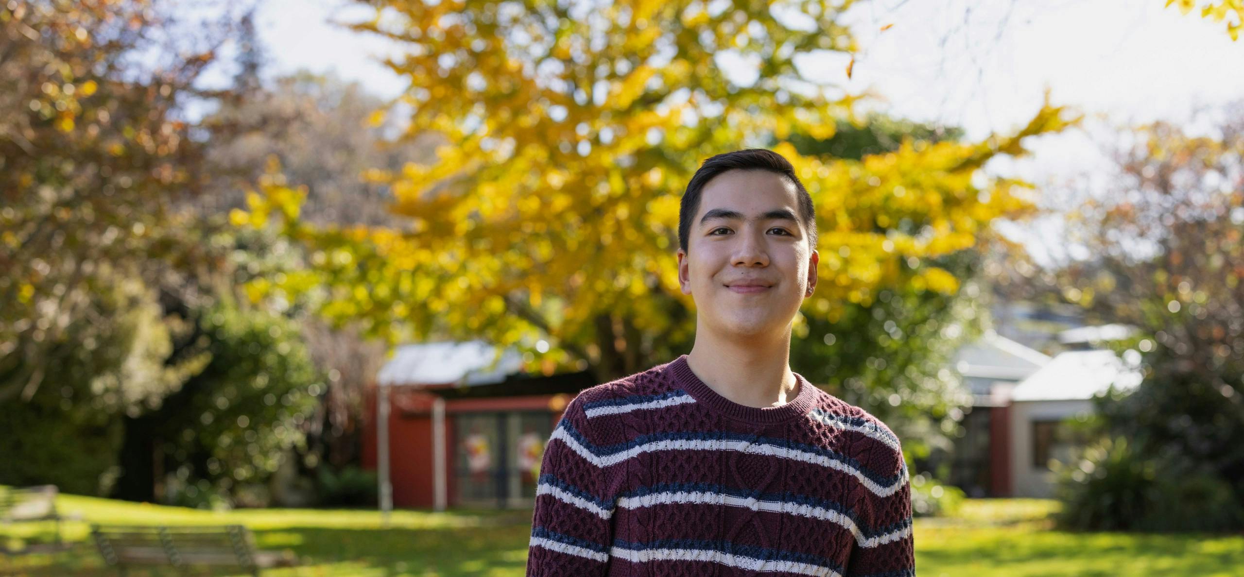 A smiling international student stands in front of a building on their campus while looking at the camera