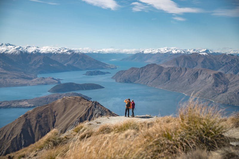 Two people walking along Roy's Peak in Wanaka and looking across the lakes and snowcapped mountains.
