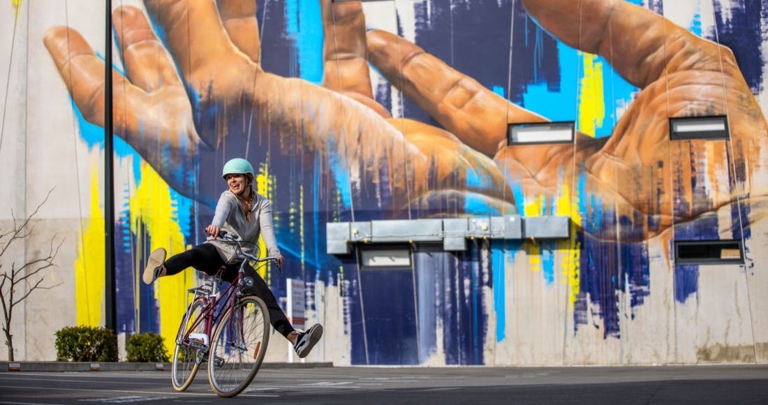 A lady riding a bike happily in front of a big wall of graffiti in Christchurch